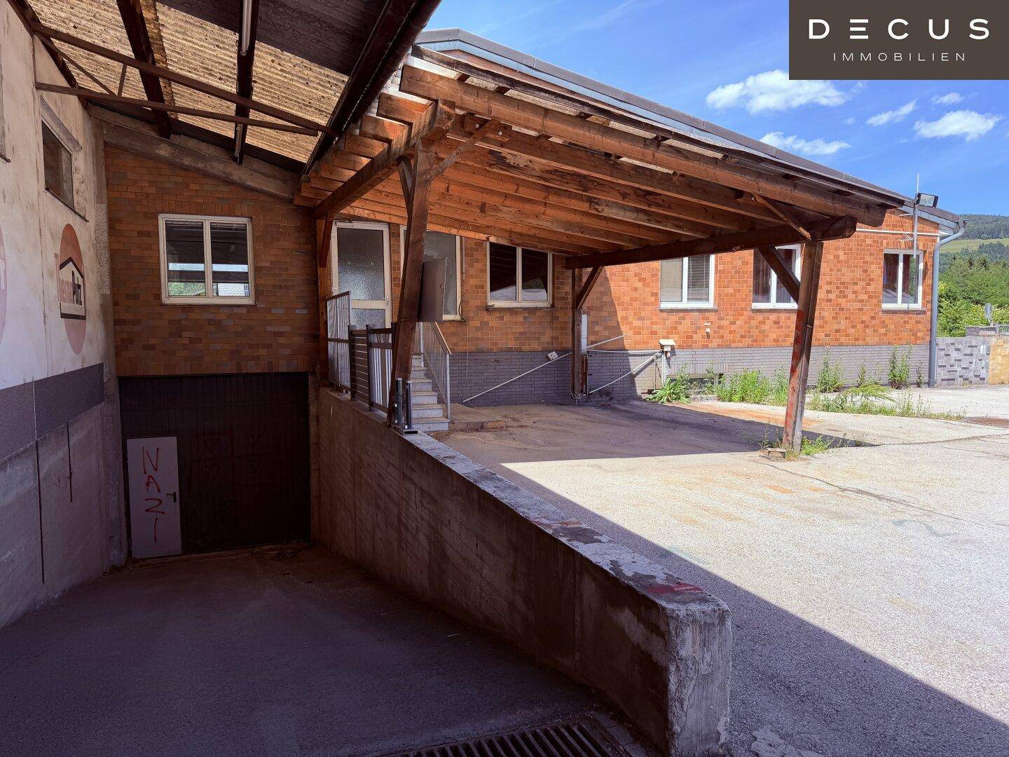 Überdachter Parkbereich oder Ladezone mit Holzbalkenkonstruktion und Ziegelgebäude im Hintergrund.