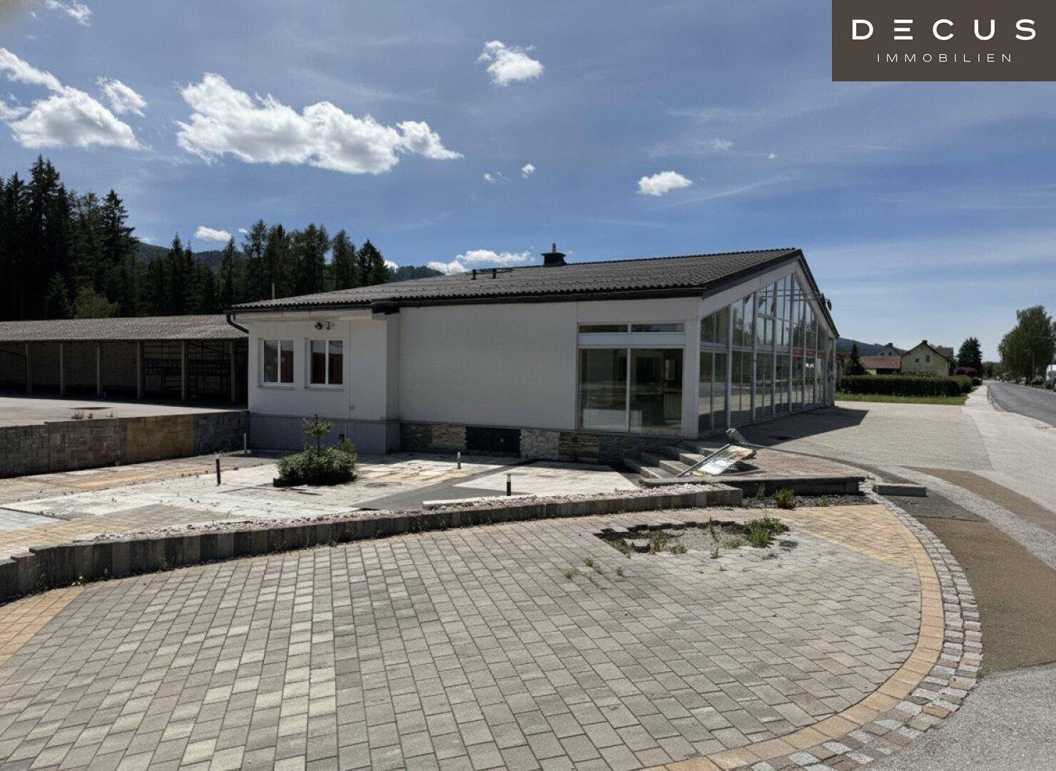 Gewerbegebäude mit großer Glasfront und gepflastertem Vorplatz, umgeben von ländlicher Landschaft.