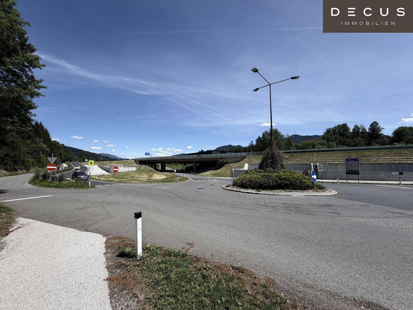 Straßenkreuzung mit Kreisverkehr und Autobahnbrücke im Hintergrund, umgeben von grüner Landschaft.