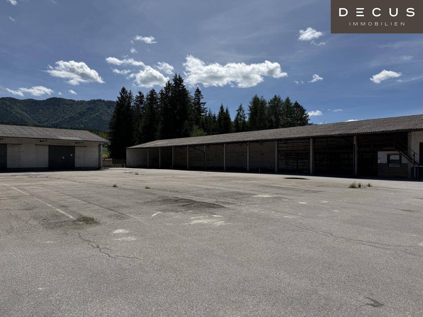 Ausgedehnte asphaltierte Fläche mit mehreren Lagerhallen im Hintergrund und bewaldeten Bergen.