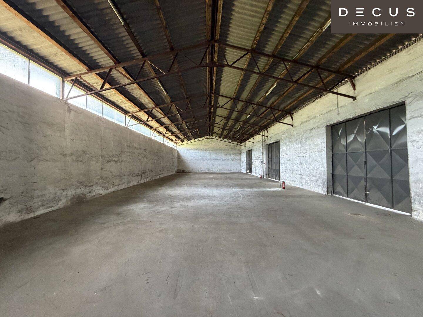 Geräumige Lagerhalle mit Betonboden, hohen Wänden und großen Metalltoren am Ende des Raumes.