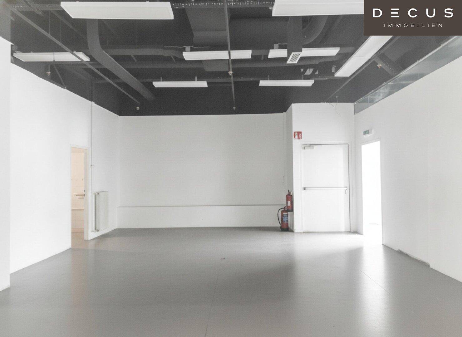 Großer, leerer Gewerberaum mit weißen Wänden, grauem Boden und sichtbaren Deckeninstallationen, bereit zur Gestaltung.