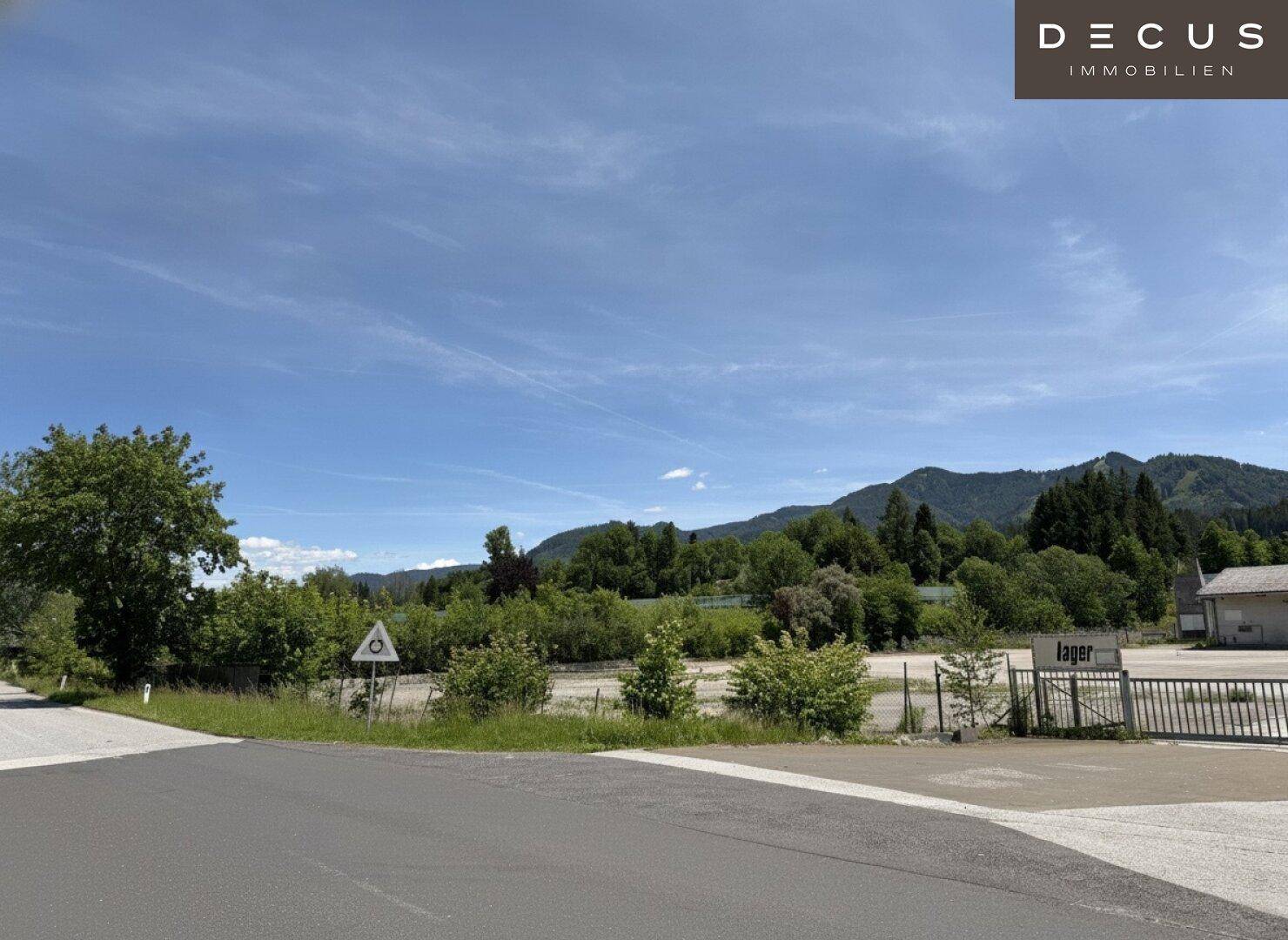 Offenes Gelände mit Bäumen und einem Schild 'Lager' im Vordergrund, umgeben von Natur und Bergen.
