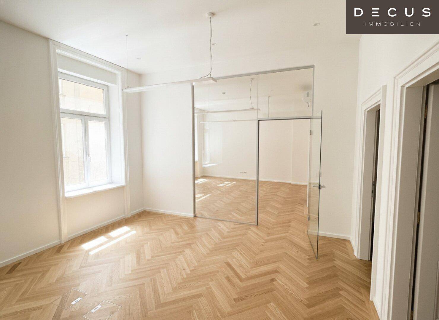 Heller Büroraum mit Fischgrätparkett, viel Tageslicht und einer modernen Glaswand.