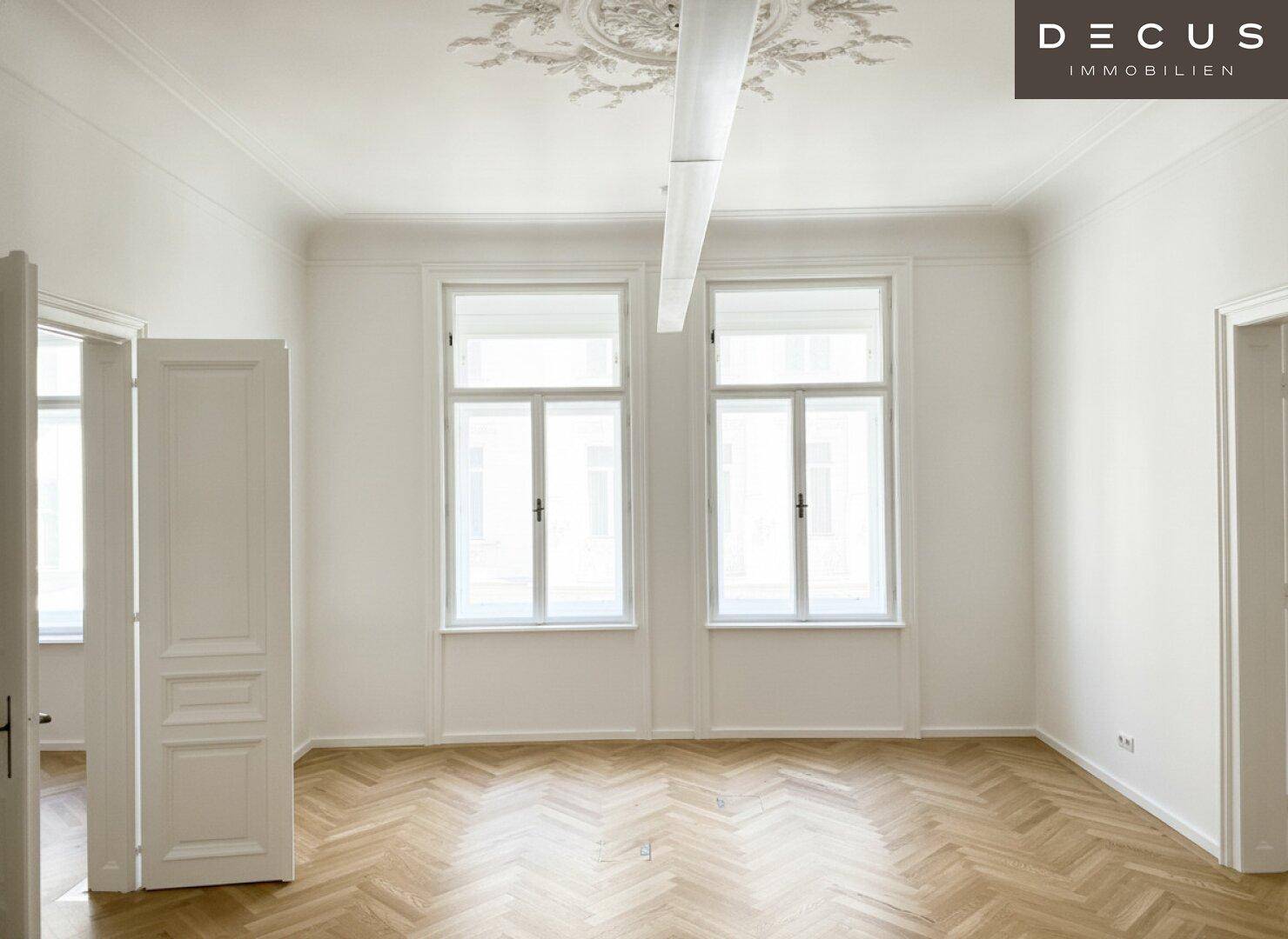 Geräumiger Raum mit Fischgrätparkett, zwei großen Fenstern und klassischer Stuckdecke.