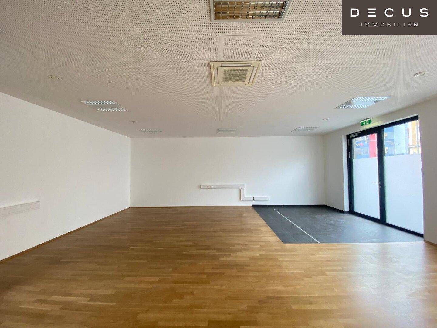 Heller, offener Bürobereich mit Holzboden und Zugang durch eine Glastür, ausgestattet mit Deckenbeleuchtung.