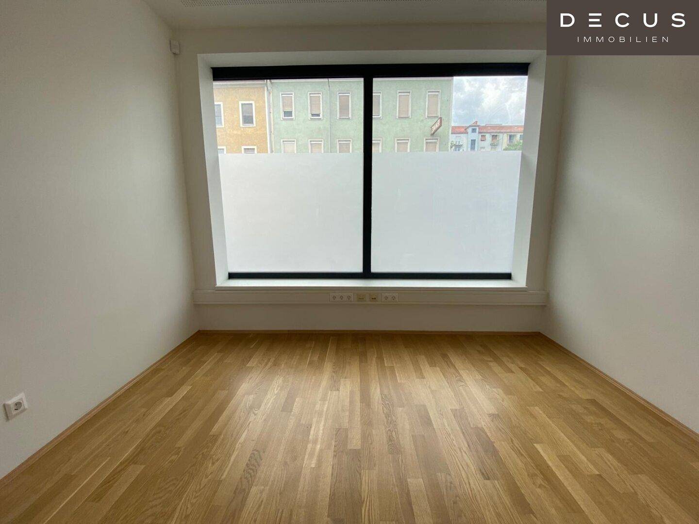 Leerer Raum mit Holzboden und einem großen Fenster, das einen Blick auf gegenüberliegende Gebäude bietet.