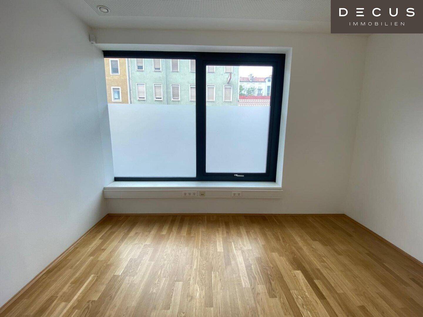 Heller Raum mit Holzboden und einem großen Fenster, das eine Aussicht auf die städtische Umgebung ermöglicht.