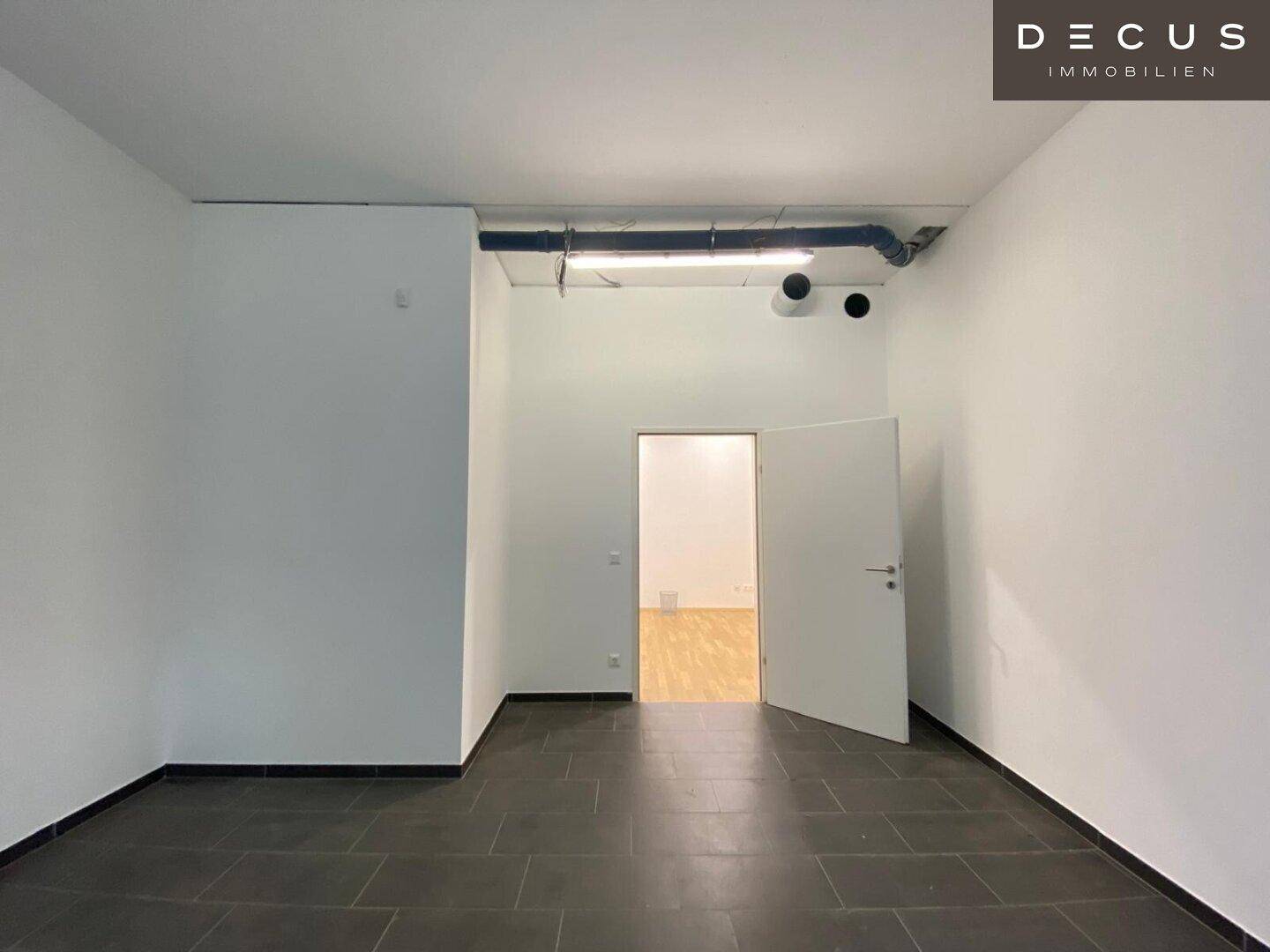 Raum mit dunklem Fliesenboden und sichtbaren Leitungen an der Decke, mit Zugang zu einem weiteren Bereich.