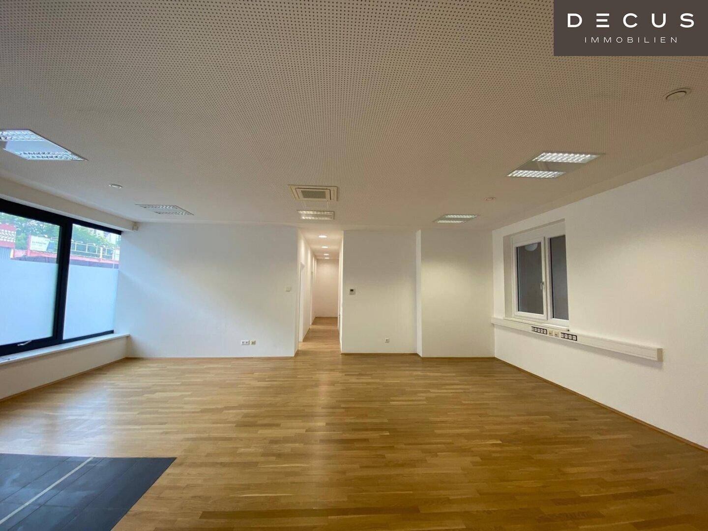 Großer, heller Büroraum mit Holzboden und mehreren Fenstern, die für reichlich Tageslicht sorgen.