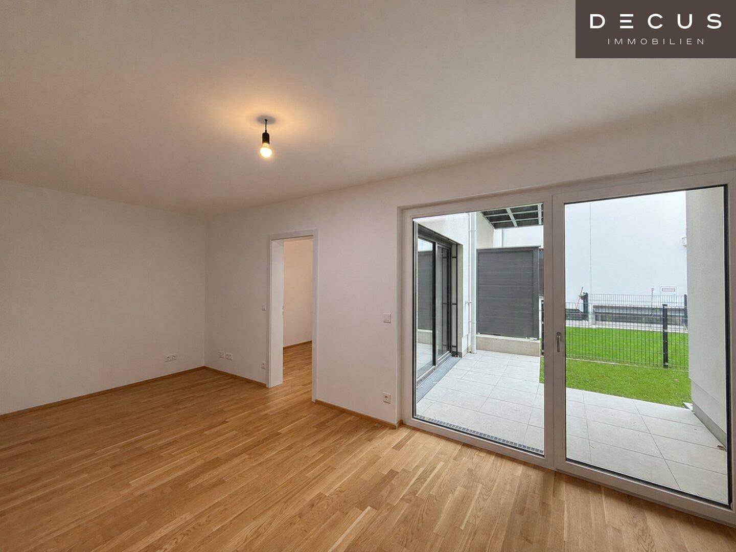 Heller Raum mit Holzboden und breiter Glasfront, die zu einer gefliesten Terrasse und Rasenfläche führt.