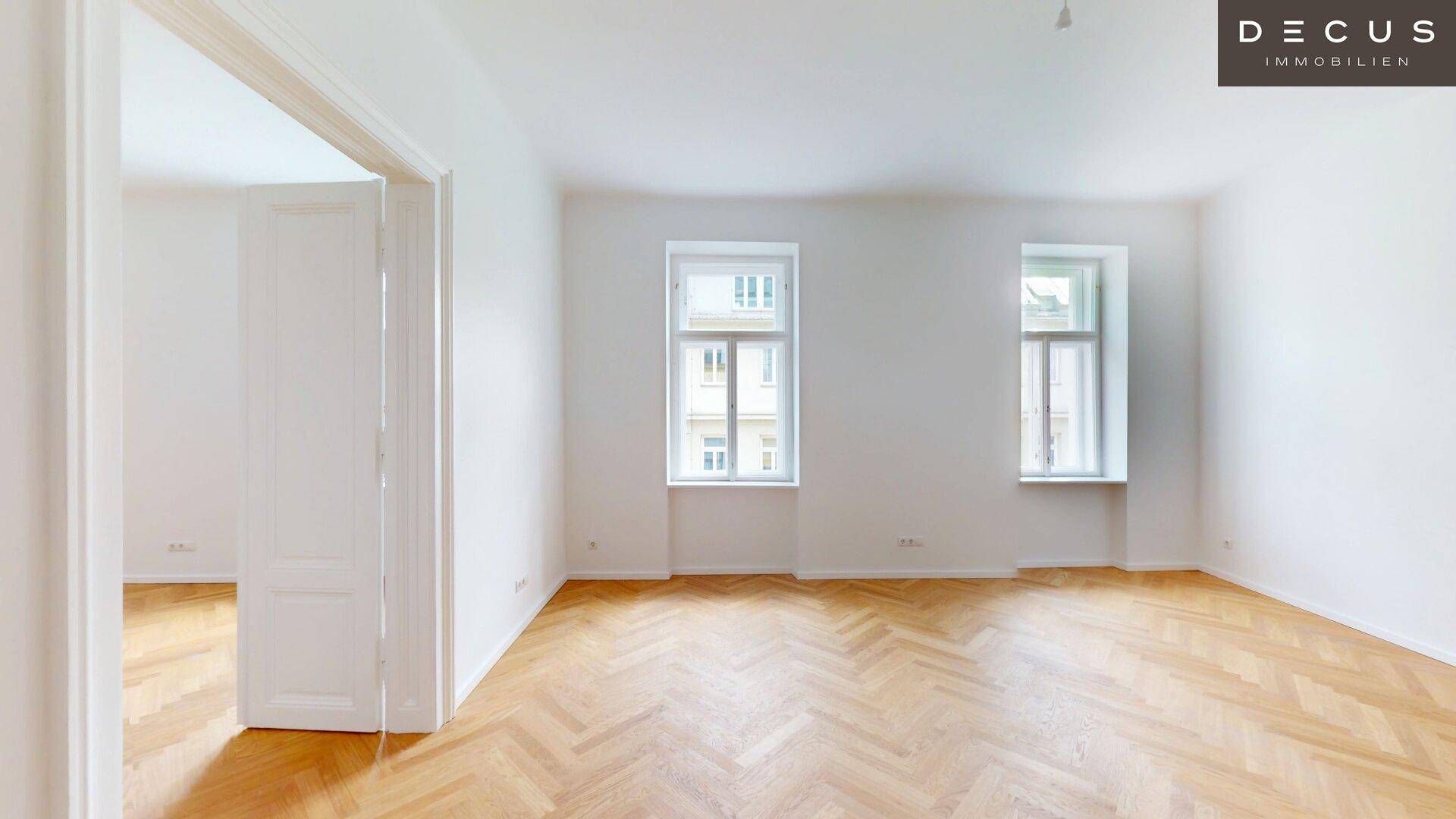 Leerer Raum mit Fischgrätparkett und zwei hohen Fenstern, der viel natürliches Licht hereinlässt.