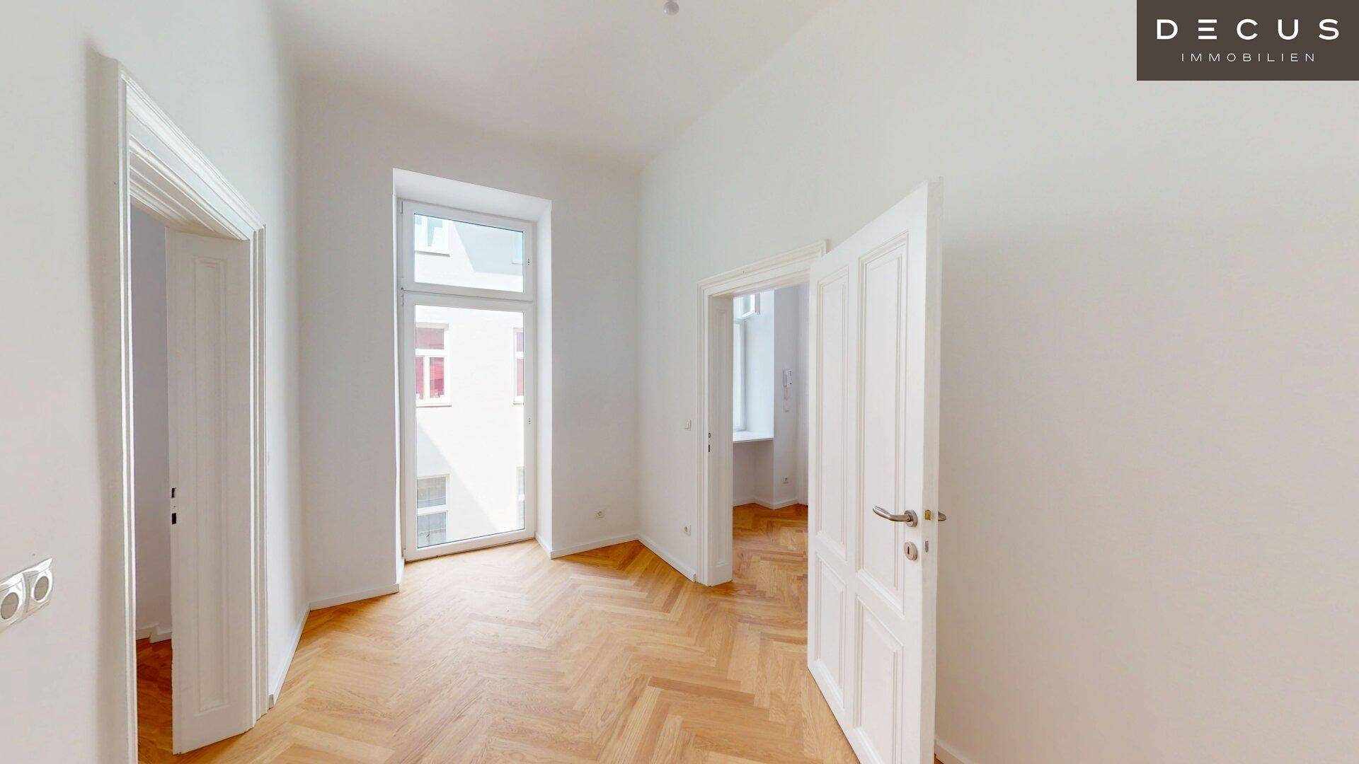 Helles Zimmer mit Fischgrätparkett und einer hohen Fenstertür, die viel Tageslicht spendet.