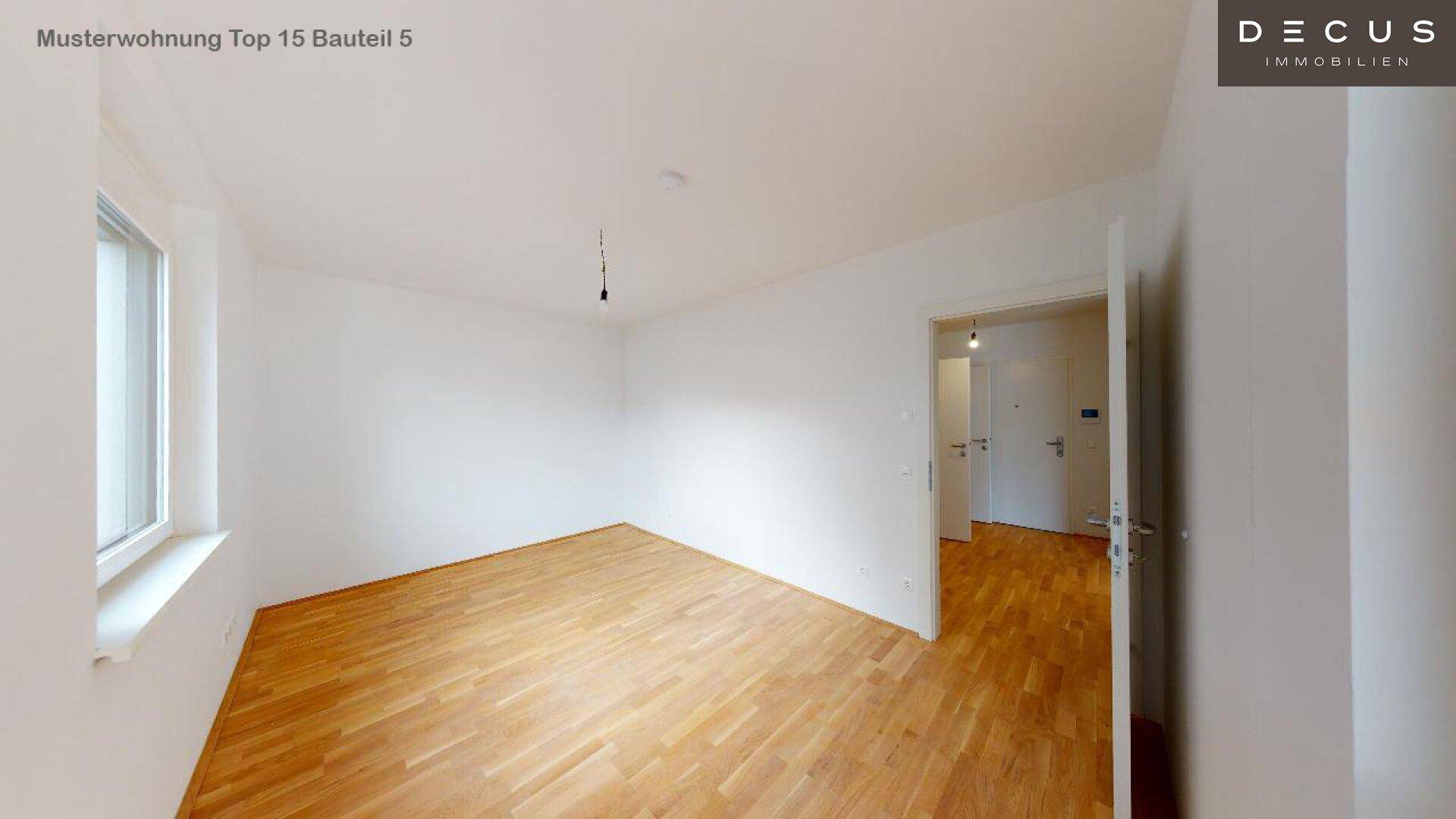 Leeres Zimmer mit hellem Holzboden, großem Fenster und Zugang zu weiteren Räumen.
