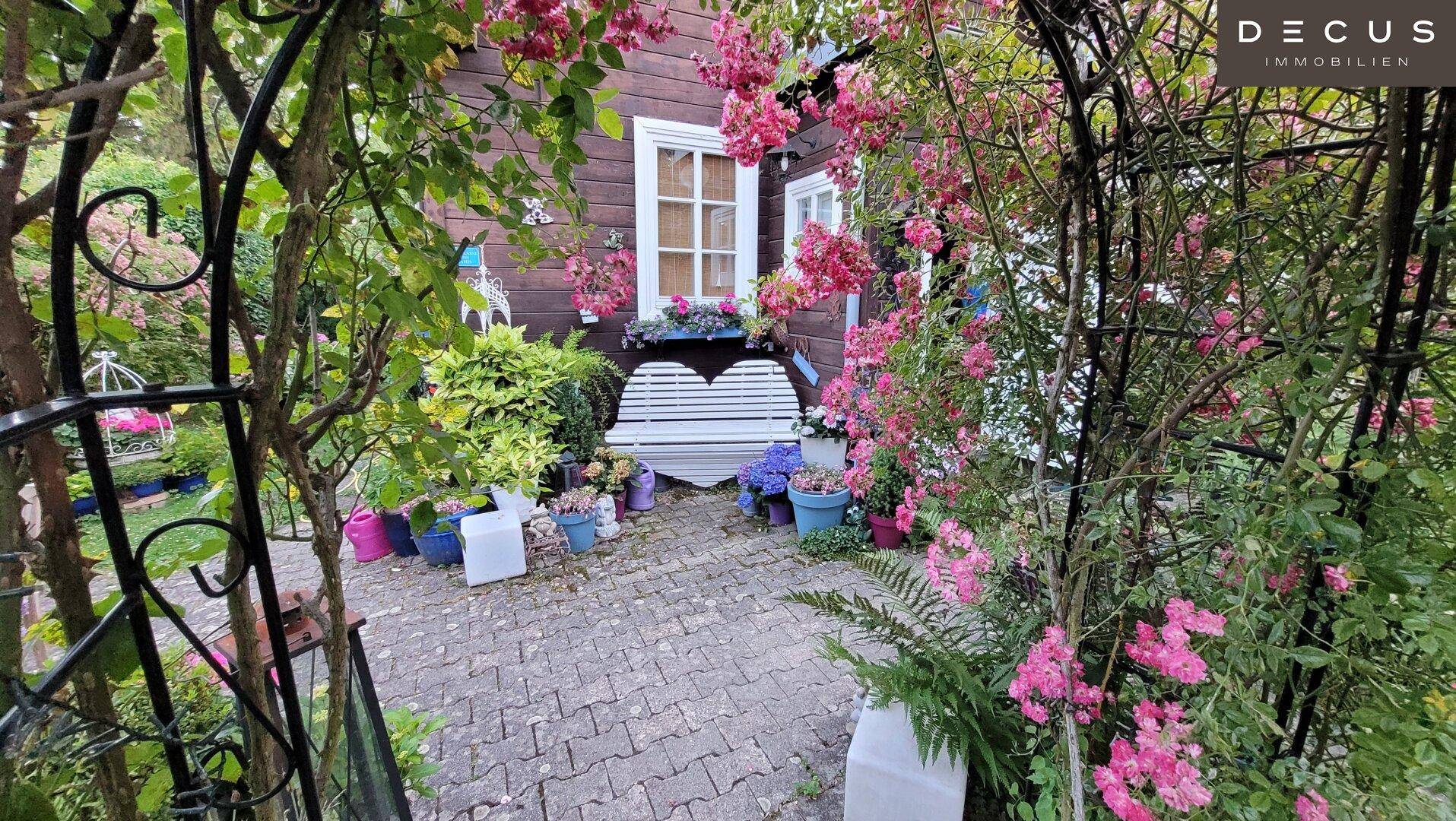 Romantischer Gartenbereich mit gepflastertem Weg, einer Herzbank und blühenden Kletterrosen am Haus.