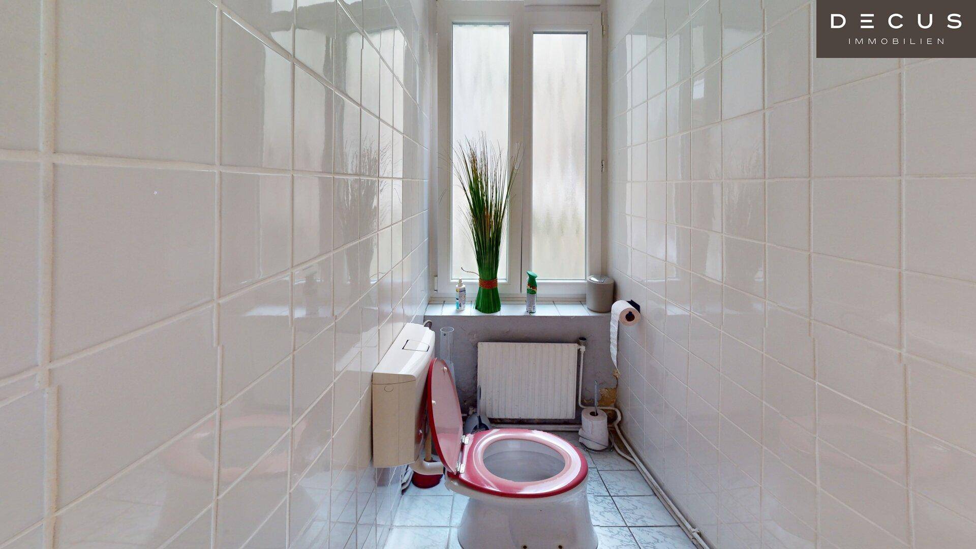 Kleine Toilette mit weißen Fliesen, Fenster und rotem Toilettensitz.