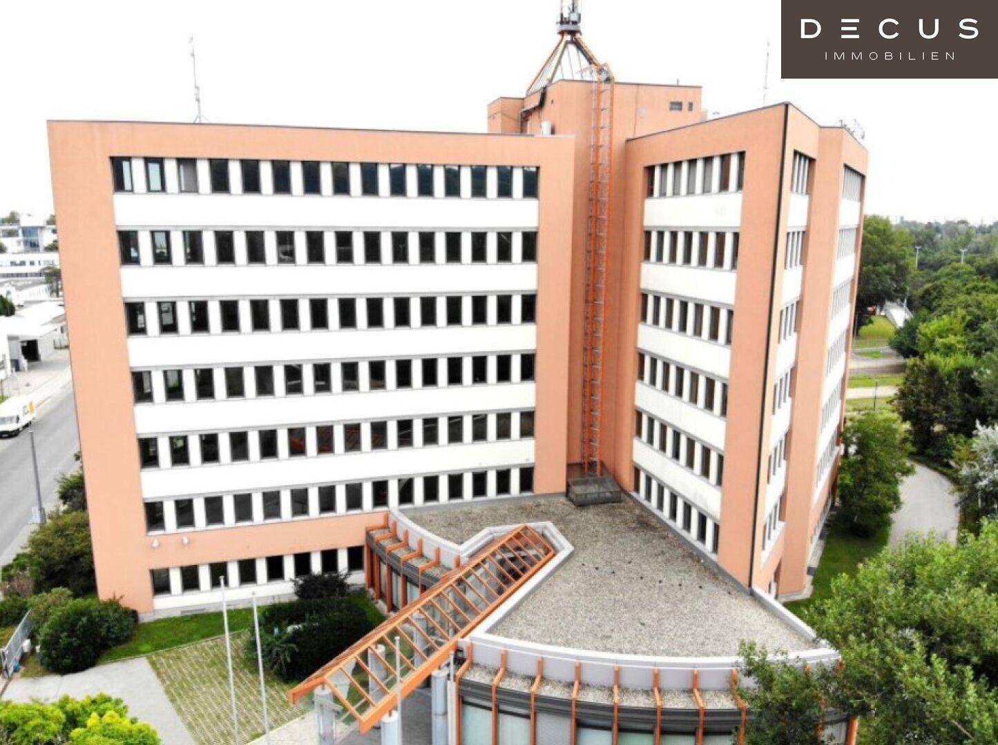 Luftaufnahme eines modernen Bürogebäudes mit orangefarbener Fassade, vielen Fenstern und umgebenden Grünflächen.