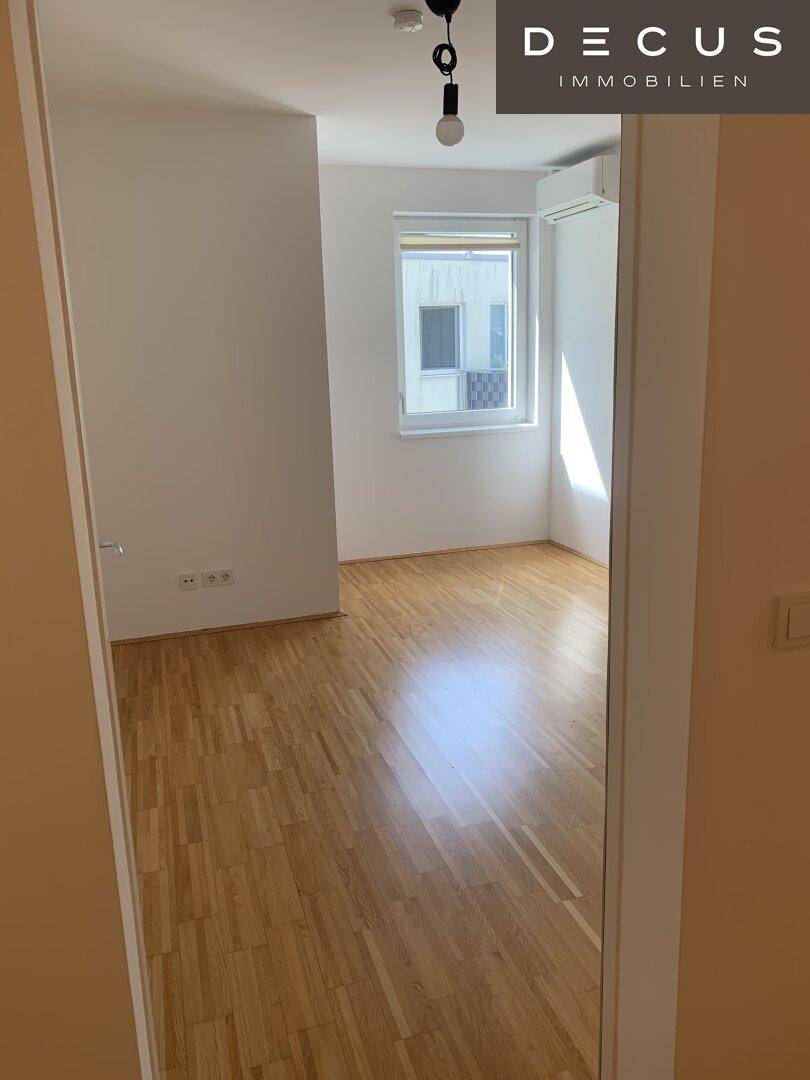 Helles, leeres Zimmer mit Parkettboden und einem Fenster, das Tageslicht hereinlässt.