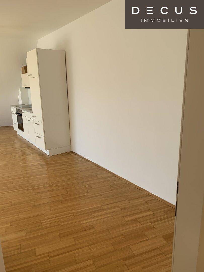 Kleine, eingebaute Küchenzeile mit weißen Schränken und Parkettboden in einem leeren Raum.