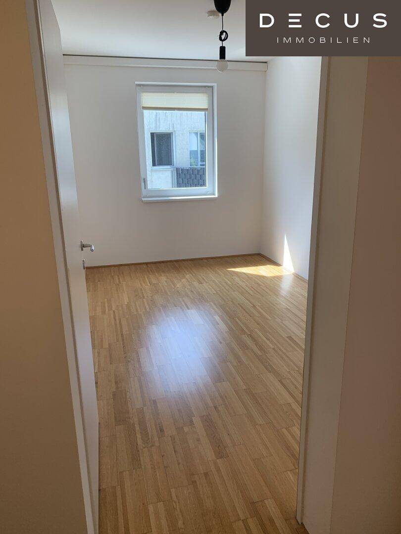 Leeres Zimmer mit hellem Parkettboden und einem Fenster, das viel natürliches Licht spendet.