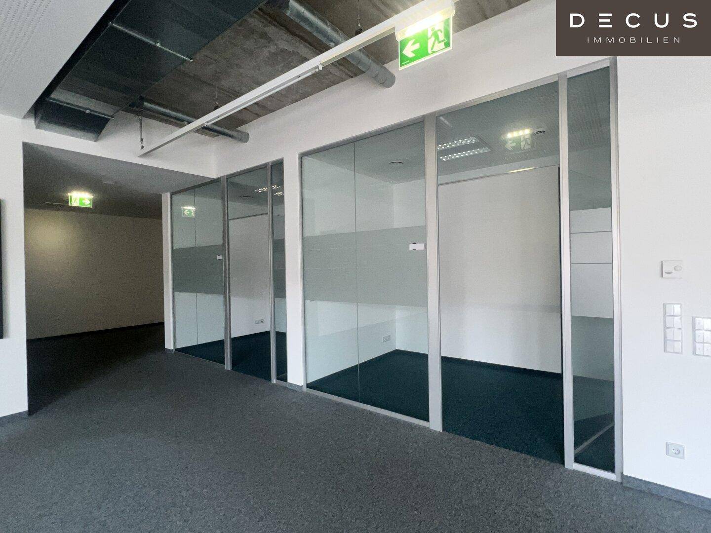 Moderner Bürogang mit Glaswänden, die separate Büros abtrennen, und sichtbaren Lüftungsrohren.