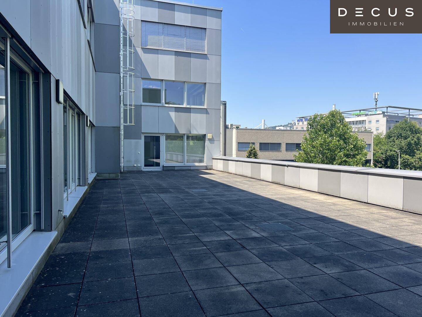 Große, gepflasterte Dachterrasse mit Blick auf das moderne Bürogebäude und den blauen Himmel.
