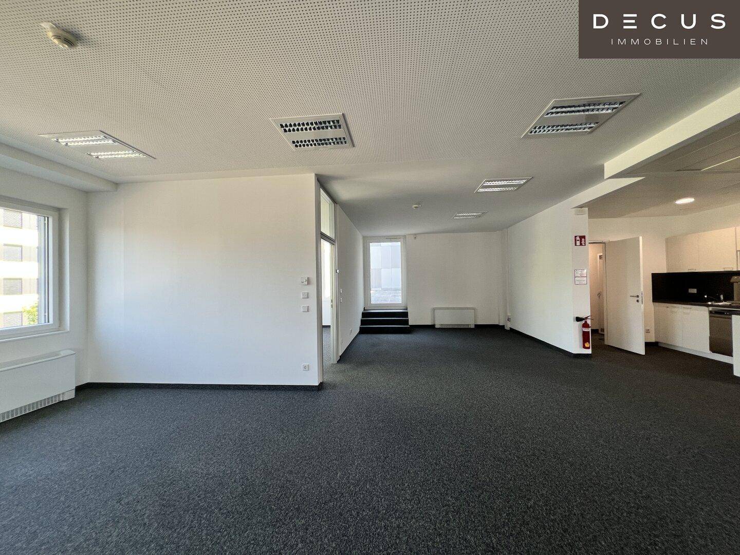 Großes, leeres Büro mit Teppichboden, weißen Wänden und einem kleinen Küchenbereich im Hintergrund.