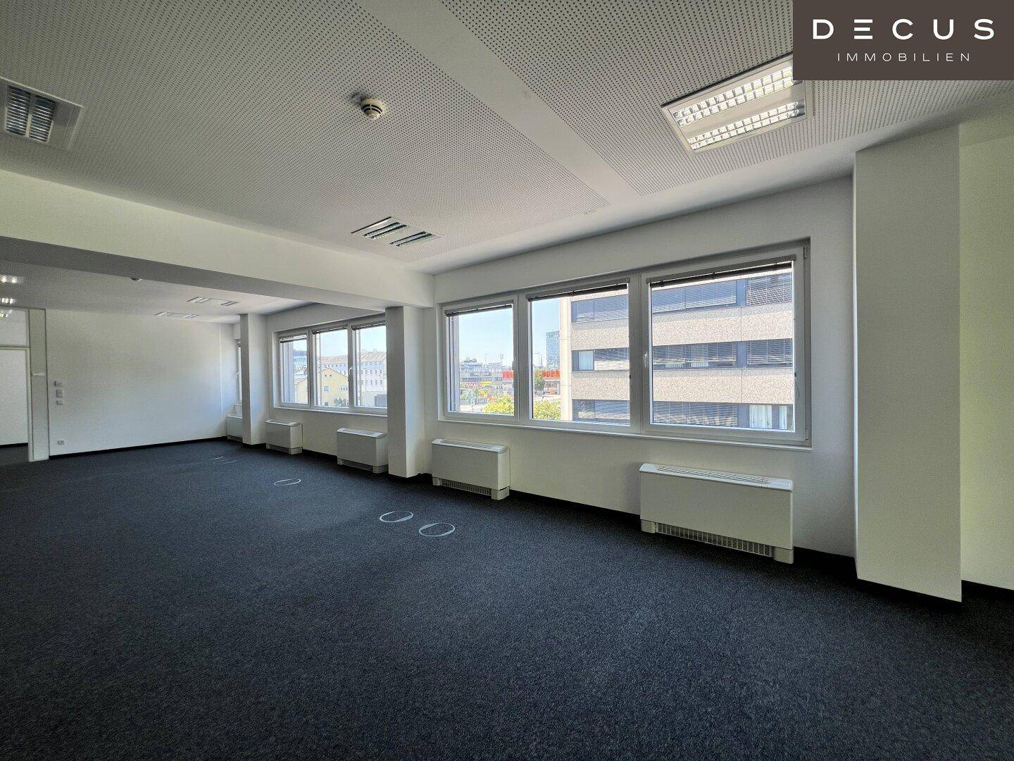 Heller Büroraum mit Teppichboden und einer Fensterfront, die einen Blick auf umliegende Gebäude bietet.