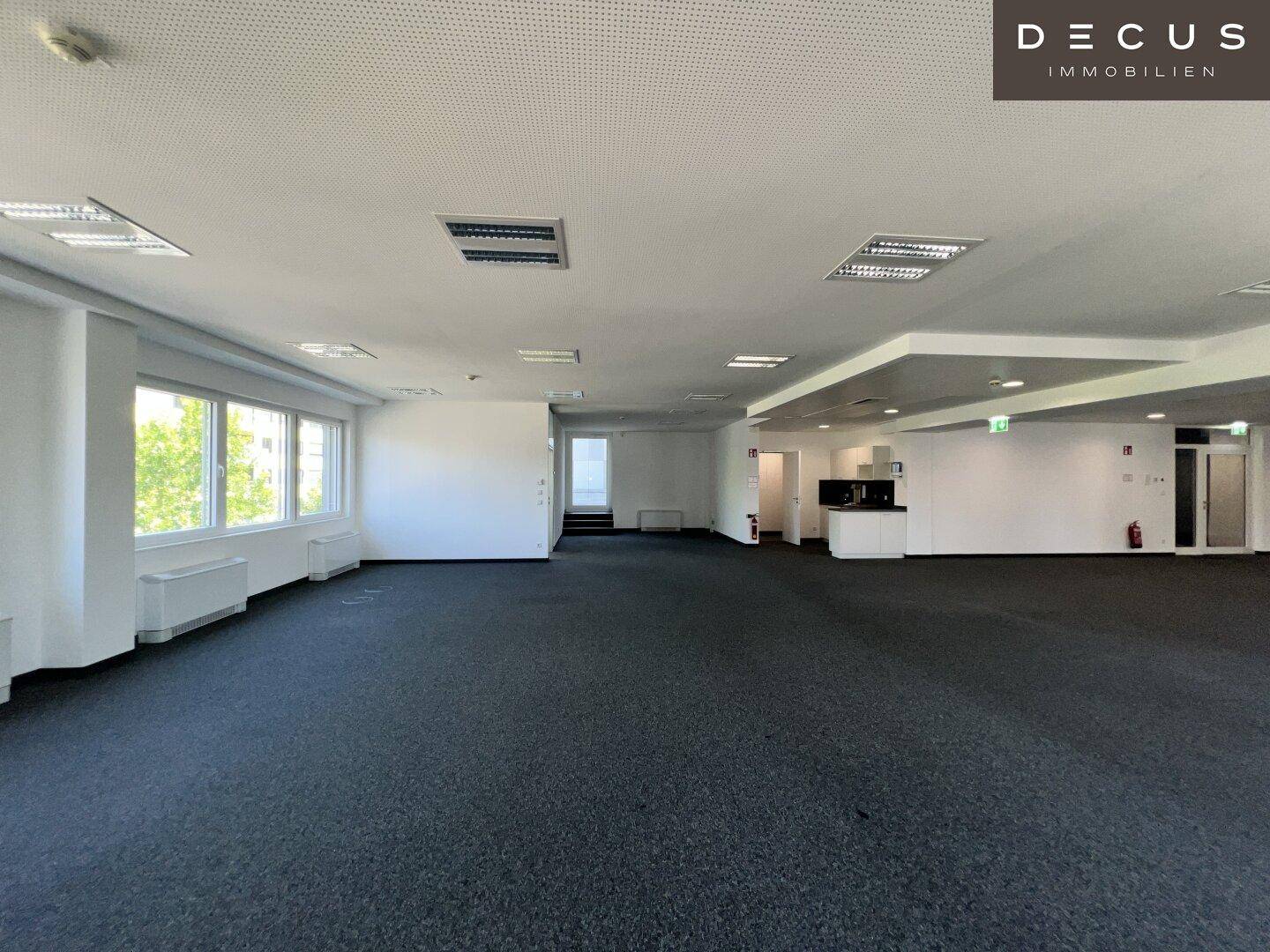 Weitläufiges, leeres Büro mit Teppichboden, vielen Fenstern und einem kleinen Küchenbereich.