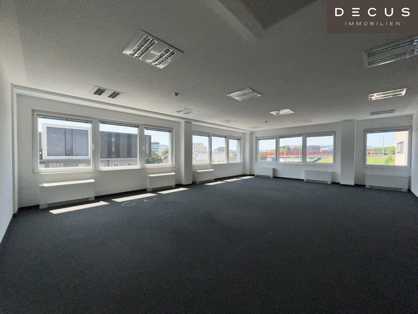Leerer Büroraum mit Teppichboden und einer langen Fensterfront, die viel Helligkeit spendet.