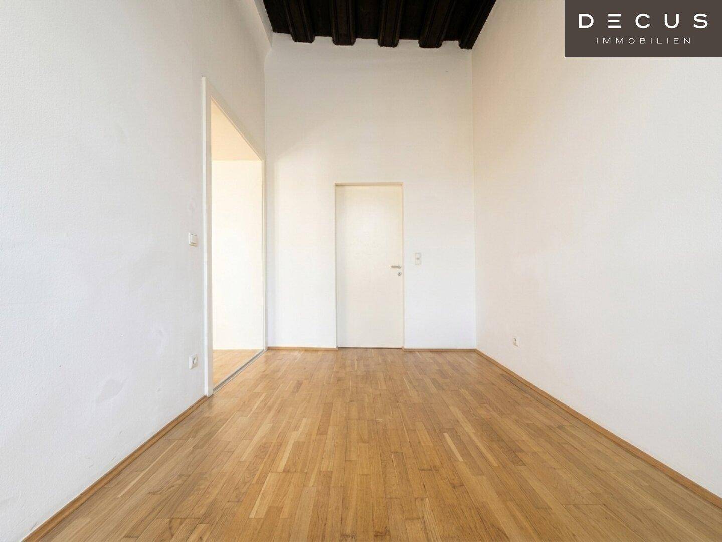 Leerer Raum mit weißen Wänden, Parkettboden und sichtbaren dunklen Holzdeckenbalken.