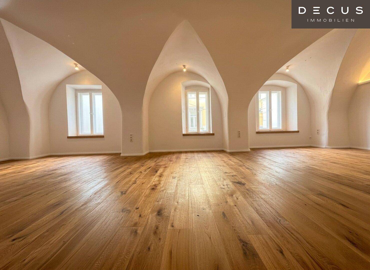Großer, heller Raum mit mehreren Fenstern, Gewölbedecken und warmem Holzboden, der viel Tageslicht hereinlässt.
