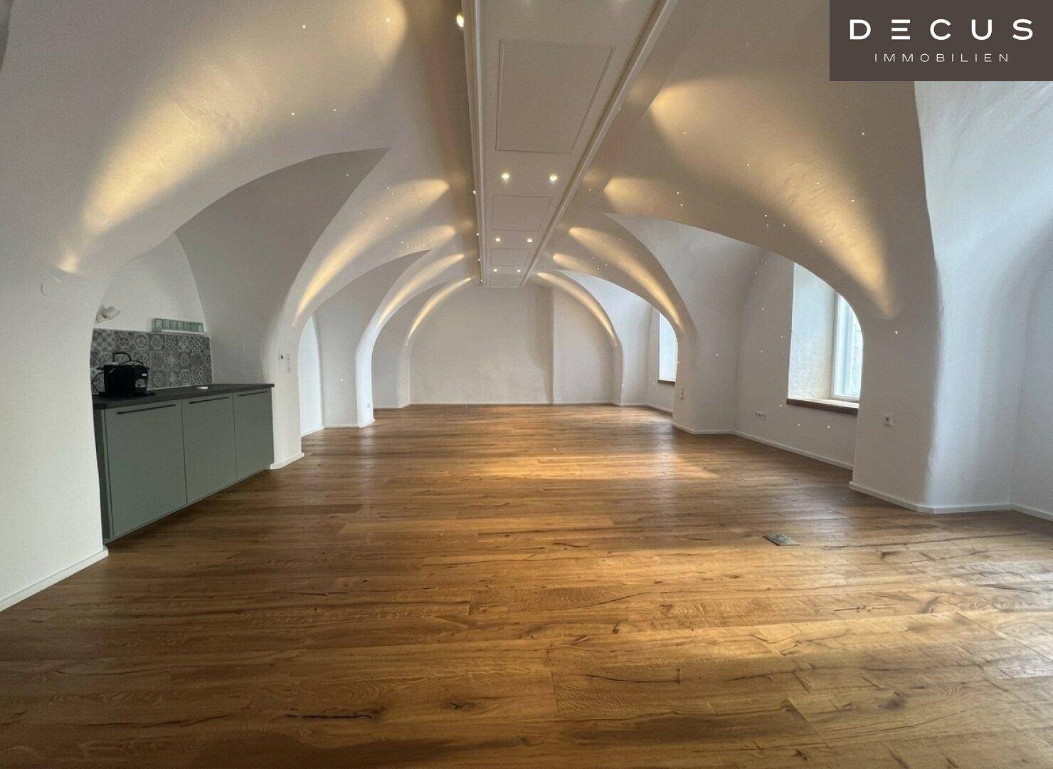 Heller Raum mit Gewölbedecken und Holzboden, ausgestattet mit einer modernen Küchenzeile und indirekter Beleuchtung.