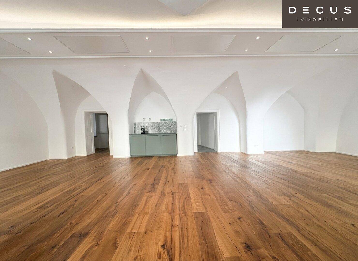 Weitläufiger Raum mit markanten Gewölbedecken und glänzendem Holzboden, inklusive einer integrierten Küchenzeile.