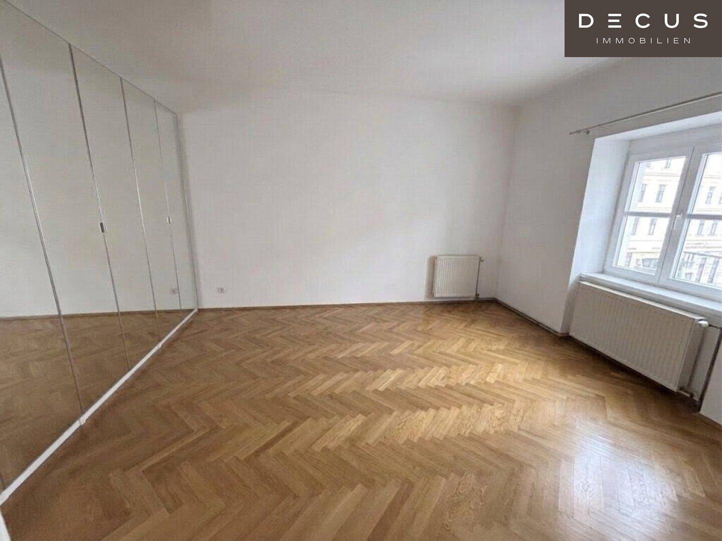 Schlafzimmer mit Fischgrätparkett, Spiegelschrank und großem Fenster.
