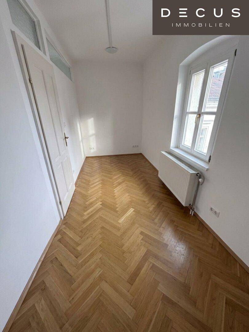 Helles Schlafzimmer mit Fischgrätparkett, weißer Tür und einem Fenster.