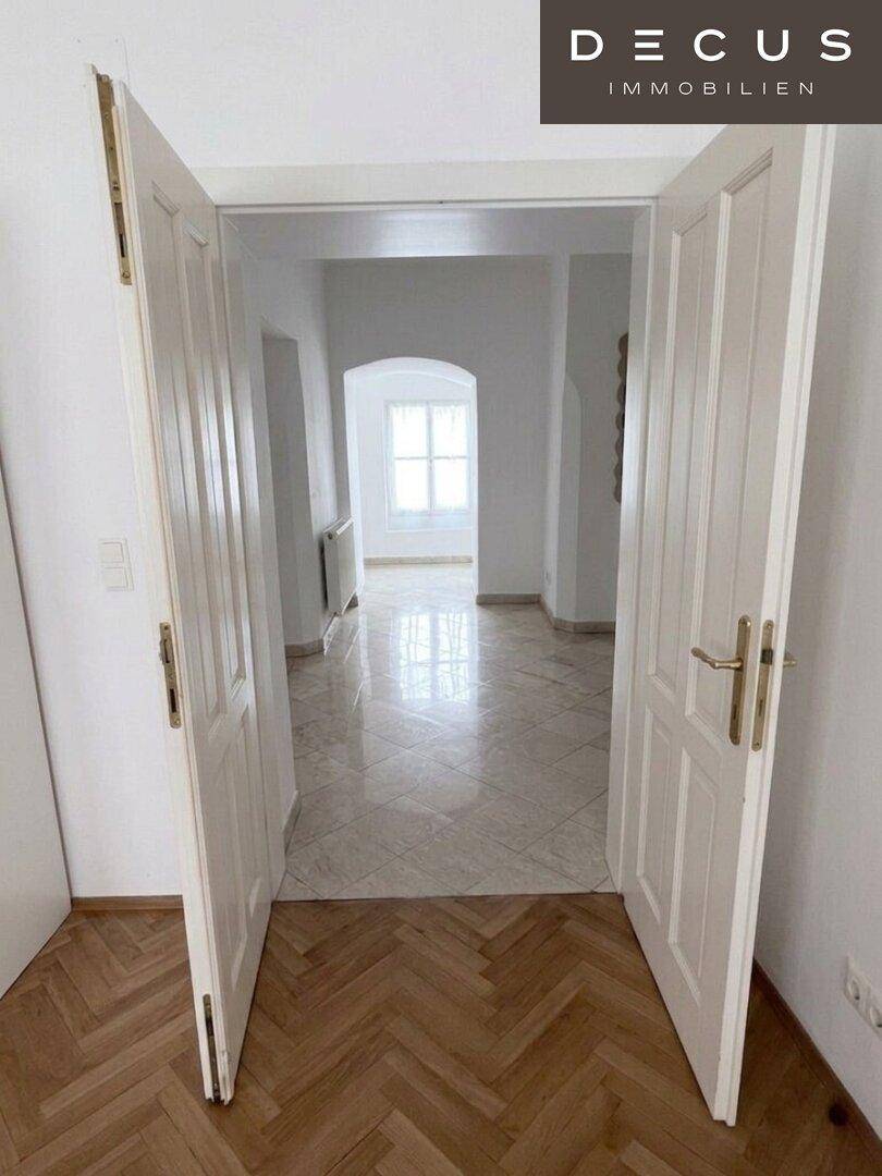 Heller Flur mit Fischgrätparkett und Blick durch eine offene Tür in einen weiteren Raum.