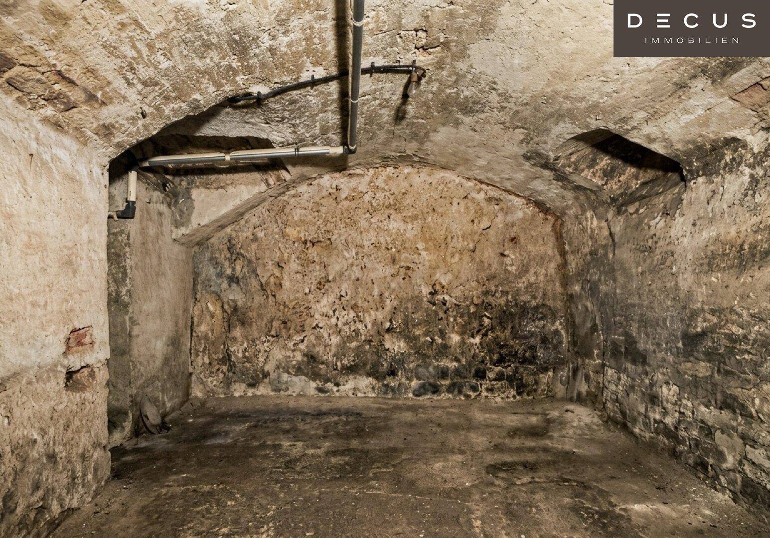Gewölbter Kellerraum mit rauen Wänden, sichtbaren Rohren und einfacher Beleuchtung.