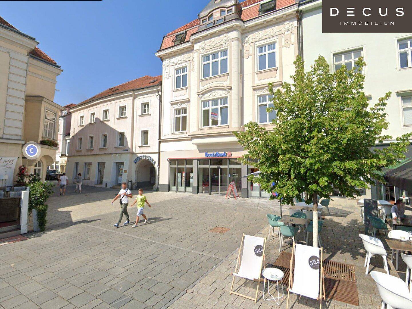Historisches Gebäude an einem belebten Stadtplatz mit Außengastronomie und Passanten.