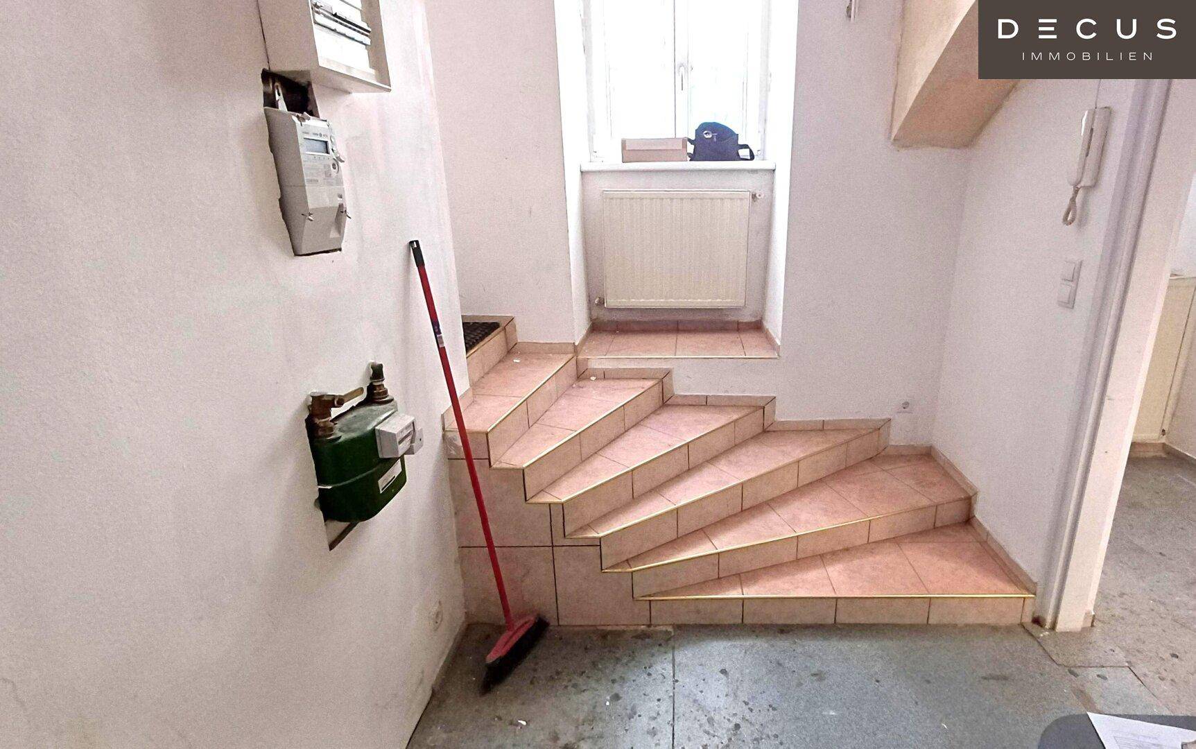 Eingangsbereich mit gefliester Treppe, Fenster, Heizkörper und Zählern an der Wand, führt in obere Etagen.