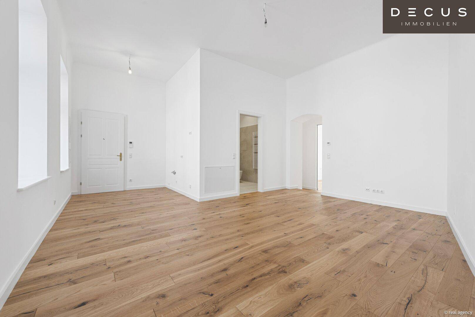 Großer, heller Raum mit Holzboden, hohen Decken und Zugang zu einem Badezimmer.