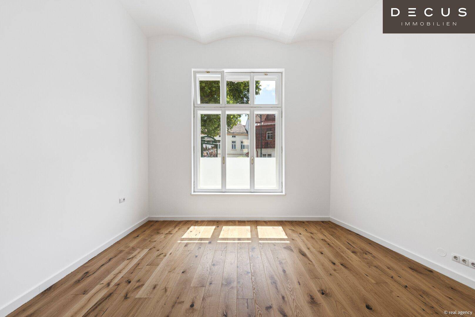 Heller Raum mit Holzboden und großem Fenster, das viel Tageslicht hereinlässt.