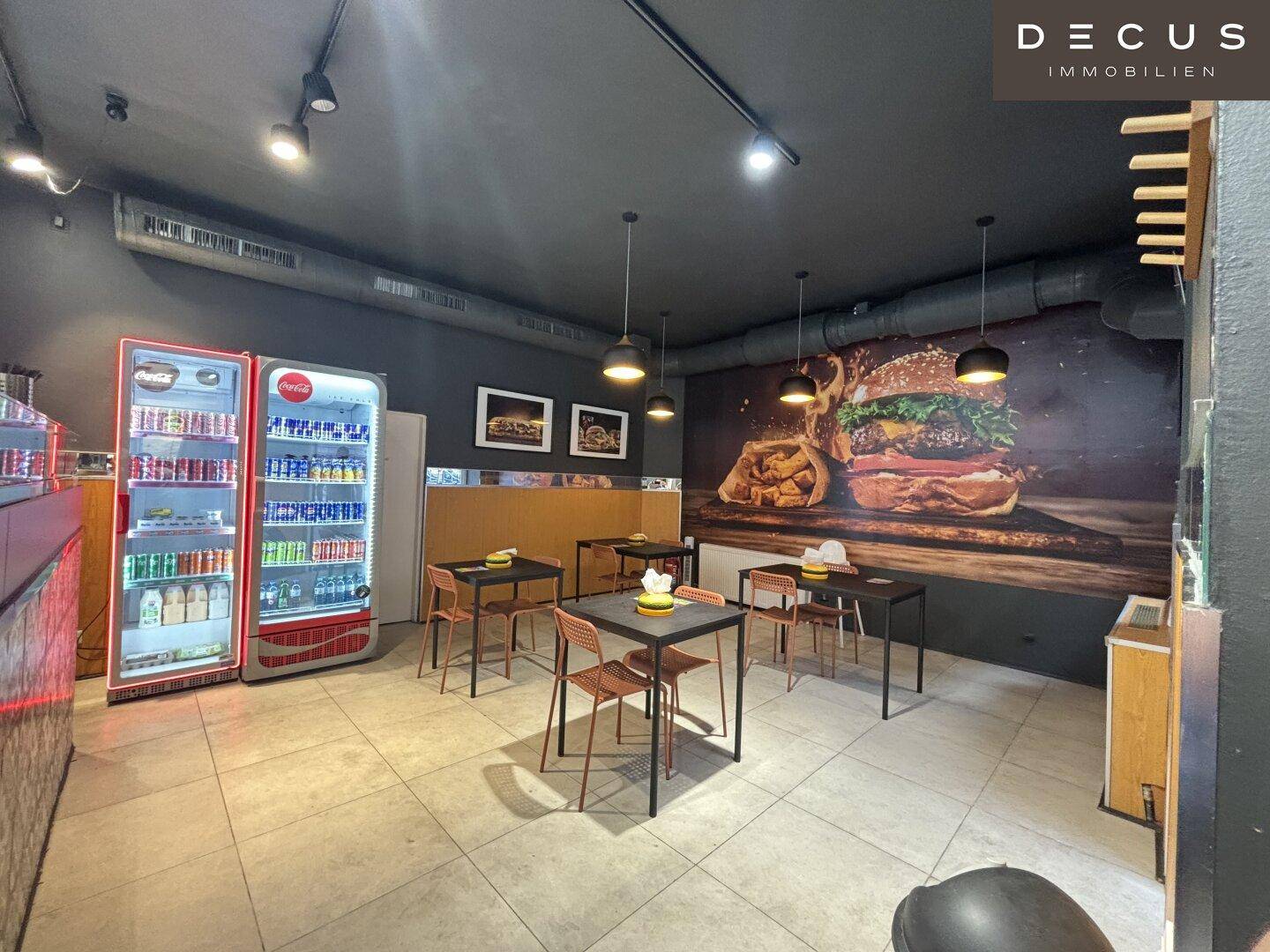 Gastraum eines Schnellrestaurants mit Wandbild eines Burgers und Getränkekühlschrank.