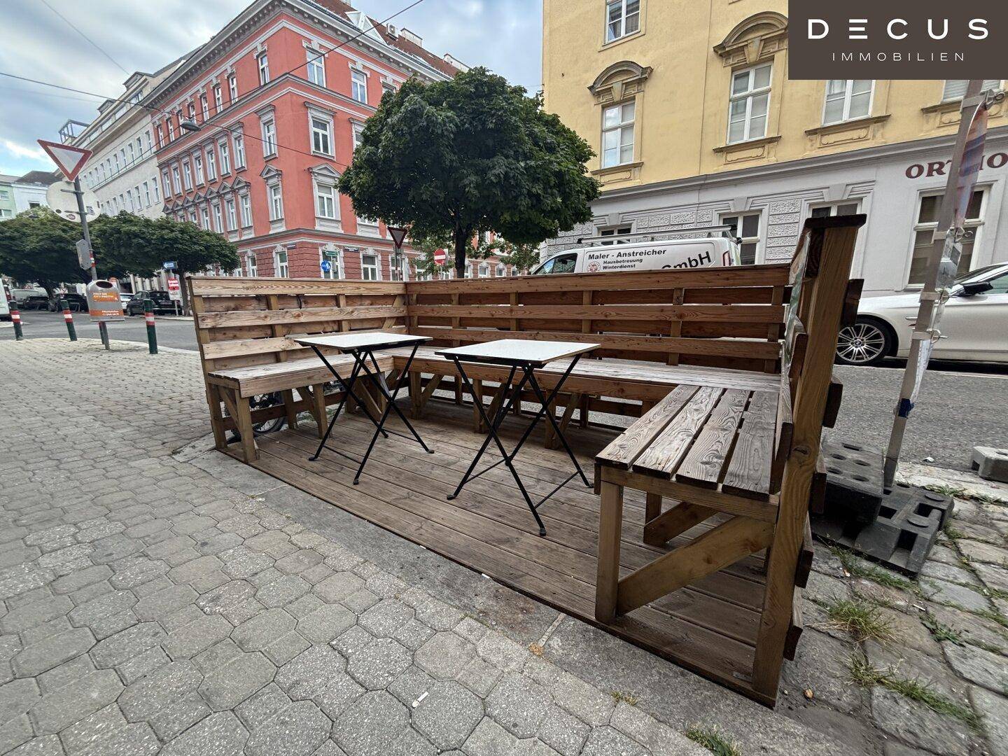 Außenbereich des Restaurants mit Holzbänken und Tischen auf einem erhöhten Podest am Bürgersteig.