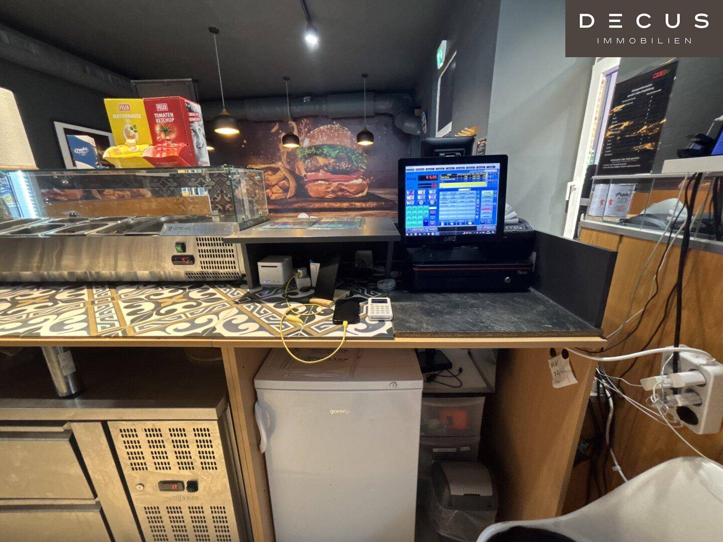 Kassenbereich mit Computer, Monitor und kleinem Kühlschrank unter der Theke.