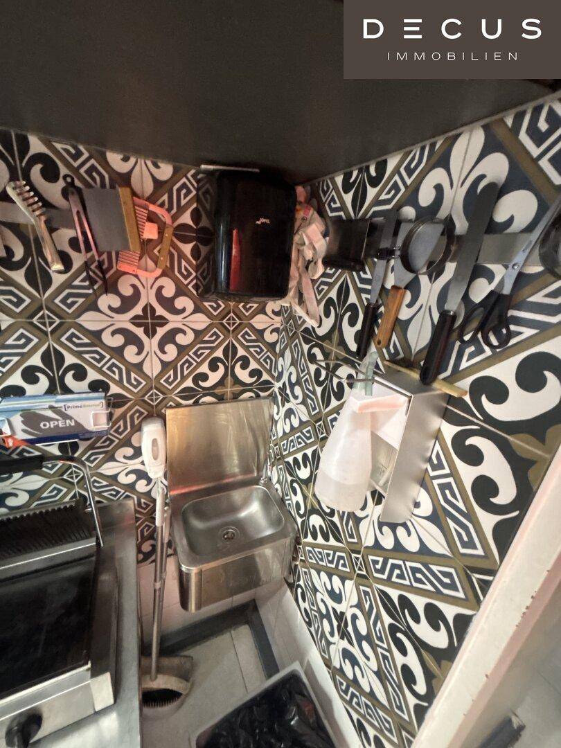 Küchenecke mit kleinem Edelstahlwaschbecken und Wandfliesen im Muster.