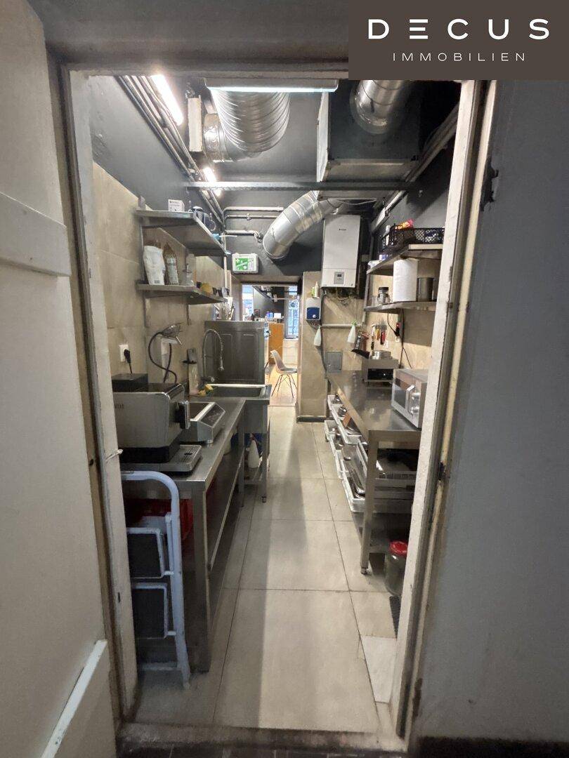Langer Küchenflur mit Edelstahltischen, Küchengeräten und sichtbaren Lüftungsrohren an der Decke.