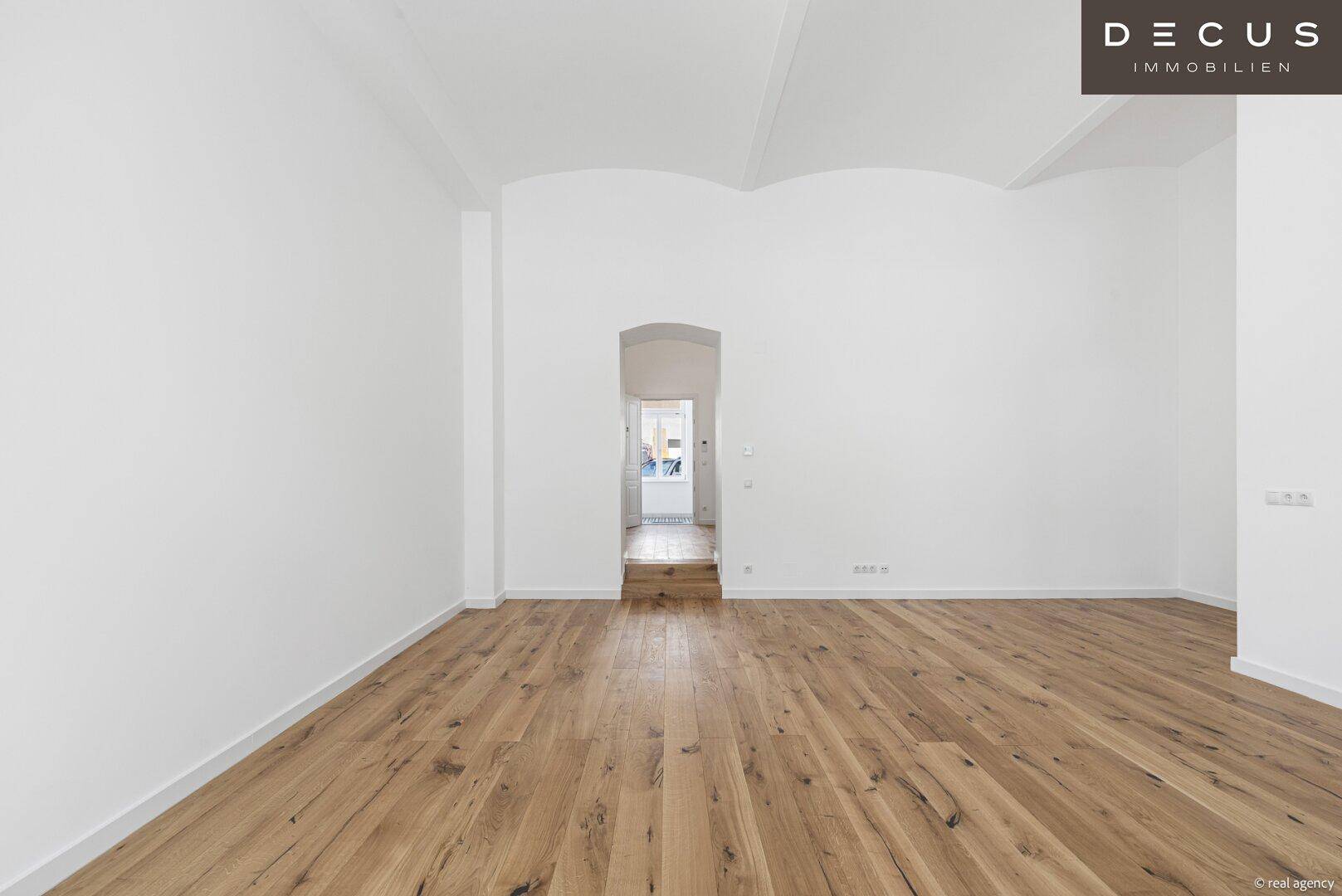 Geräumiges, leeres Wohnzimmer mit Holzboden, hohen Decken und einem Durchgang zu weiteren Räumen.