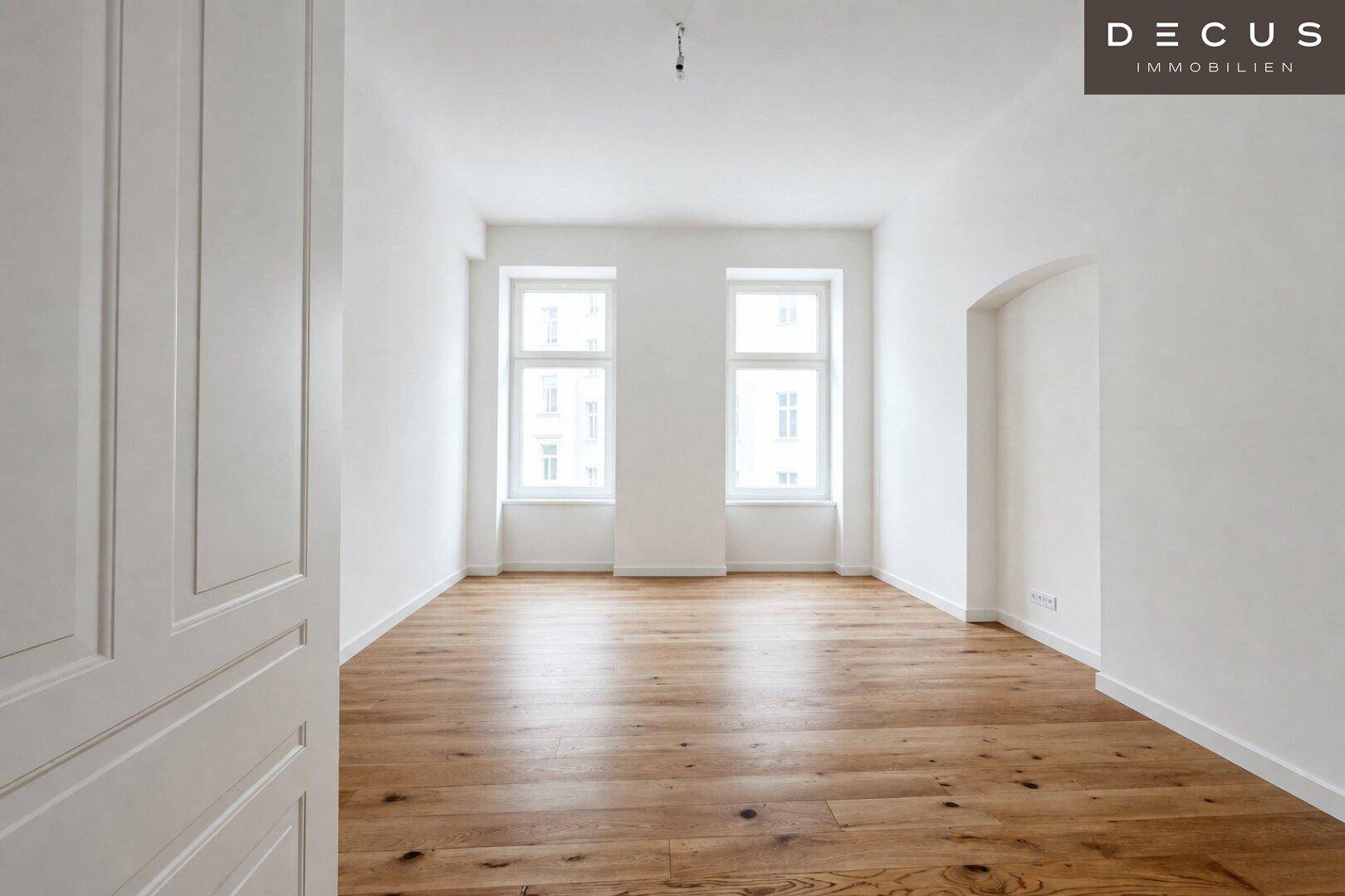 Leeres Schlafzimmer mit hellem Holzboden und zwei Fenstern, die für viel Tageslicht sorgen.