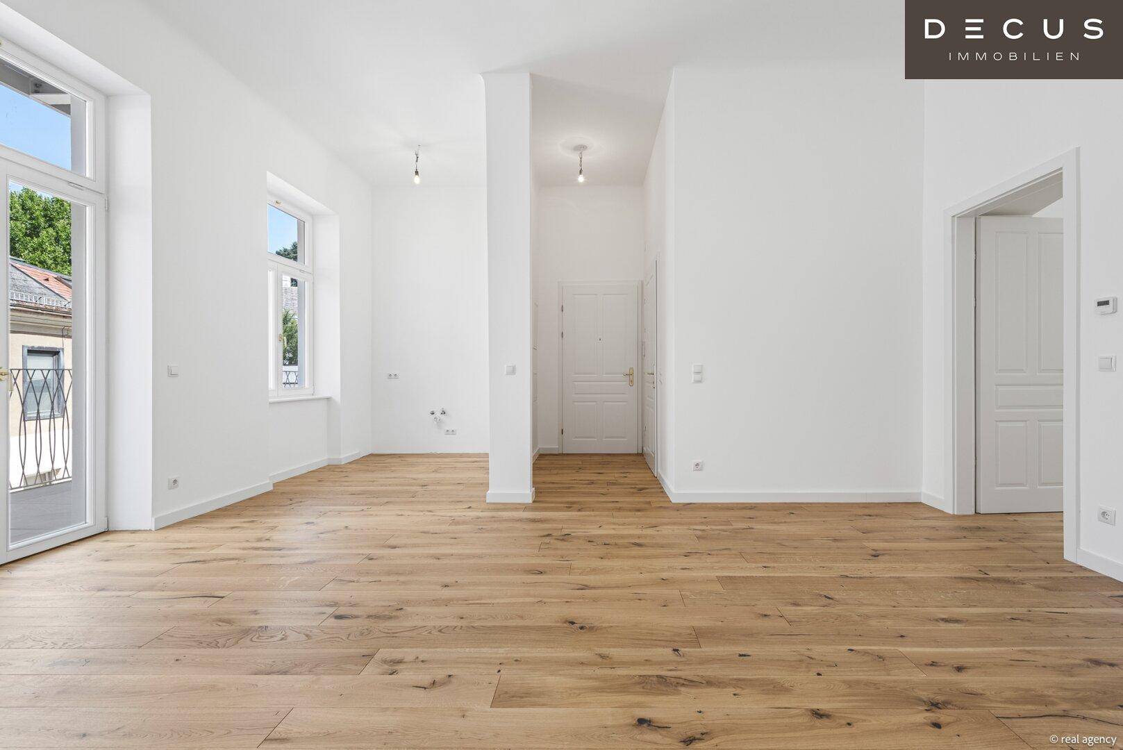 Geräumiger, heller Raum mit Holzboden, weißen Wänden und Zugang zu weiteren Zimmern.